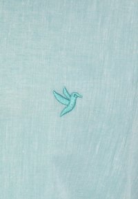 Un tejido azul claro presenta un pequeño colibrí bordado en un tono ligeramente más oscuro de azul. La textura parece tejida y suave.