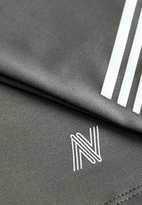 Szary materiał sportowy z pionowym prążkowaniem, z białym logo i trzema białymi paskami wzdłuż krawędzi. Gładka faktura.
