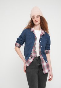 Γυναίκα με σγουρά μαλλιά φοράει ροζ σκουφάκι, μπλε πλεκτή ζακέτα πάνω από ροζ γκρι καρό πουκάμισο και μαύρο τζιν, στέκεται με το ένα χέρι στη μέση.