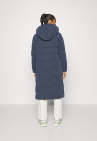 Lang marineblå dunfrakke med hætte, med et quiltet design og en let indsnævret talje. Sammen med lyse bukser og sneakers.
