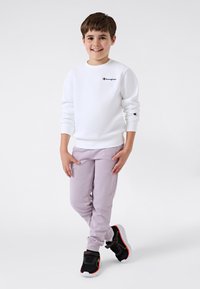 Leende pojke står med benen i kors, iförd en vit Champion-hoodie, ljuslila byxor och svarta sneakers med röda och gröna detaljer.