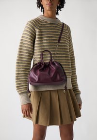 Sac en cuir violet avec un design froncé, deux poignées courtes et une longue sangle, posé sur un mannequin portant un pull rayé et une jupe.