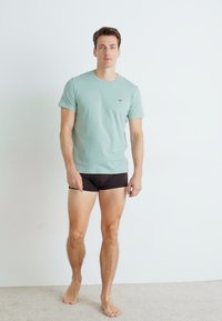 Camiseta verde claro de cuello redondo, hecha de tela suave, combinada con pantalones cortos negros ajustados. El modelo está descalzo contra un fondo liso.