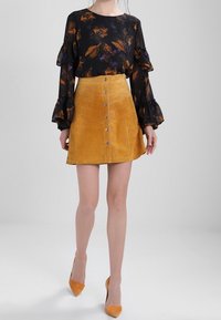 Blouse noire à fleurs avec manches à volants, associée à une jupe taille haute en daim moutarde avec des boutons sur le devant. Complété par des escarpins assortis.