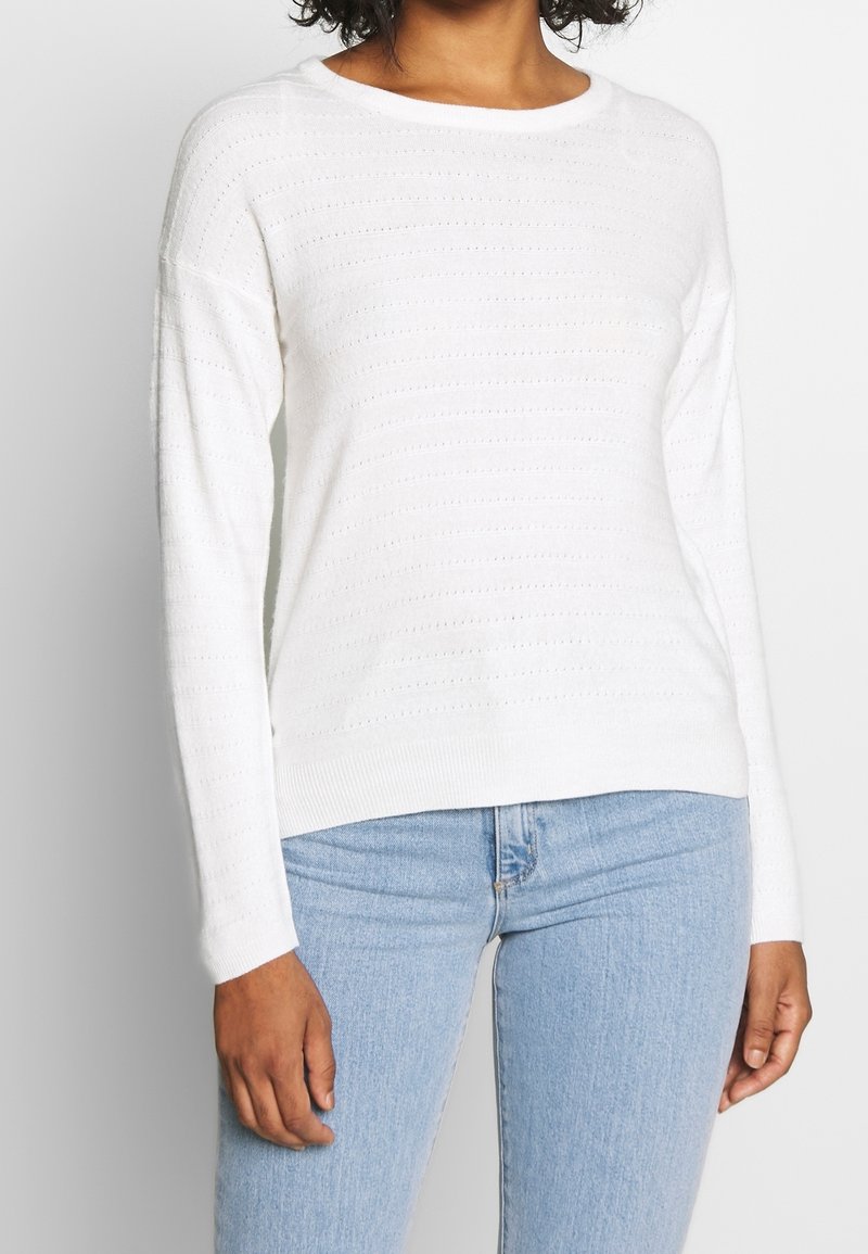 Woman wearing a white long-sleeve knit sweater with subtle horizontal perforated stripes and light blue denim jeans.