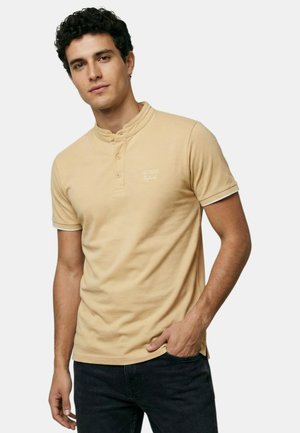 Jeune homme aux cheveux bouclés et foncés portant un t-shirt henley beige à manches courtes et un pantalon noir, debout avec une main dans la poche.