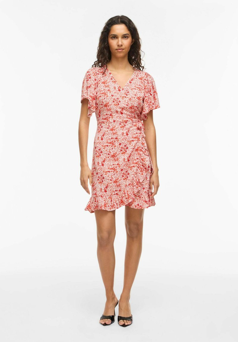 Femme debout portant une robe portefeuille fleurie rose longueur genou avec des manches courtes volantées et des talons blocs ouverts noirs sur fond blanc.