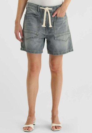 Persona che indossa pantaloncini casual grigi scoloriti con grandi tasche frontali e un cordoncino bianco, abbinati a sandali bianchi con tacco.