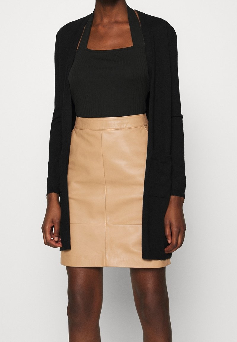 Femme portant un débardeur noir, un cardigan noir et une mini-jupe en cuir beige, debout devant un fond clair uni.