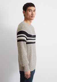 Beiger Strickpullover mit schwarzen und weißen horizontalen Streifen über der Brust. Rundhalsausschnitt, lange Ärmel und strukturiertes Material.