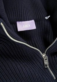 Laivastonsininen neule, jossa on vetoketju hopeisella vetimellä. Sisäisessä lapussa lukee "JJXX" vaalean lilalla tarralla. Teksturoitu kangas, jossa on ribattua pintaa.