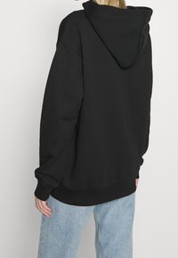 Felpa nera con cappuccio e vestibilità ampia, caratterizzata da un cappuccio grande, polsini e orlo a coste. Indossata con jeans di denim azzurro chiaro.