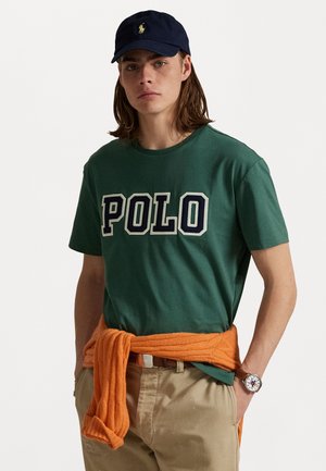 Groene katoenen T-shirt met "POLO" logo, gecombineerd met een oranje trui om de taille, beige broek en een marineblauwe pet. Witte horloge om de pols.