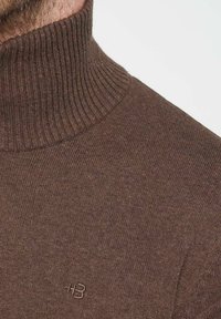 Brauner gestrickter Rollkragenspullover mit geripptem Kragen, ausgestattet mit einem dezenten gestickten Logo auf der Brust. Glatte Textur, figurschnittige Passform.