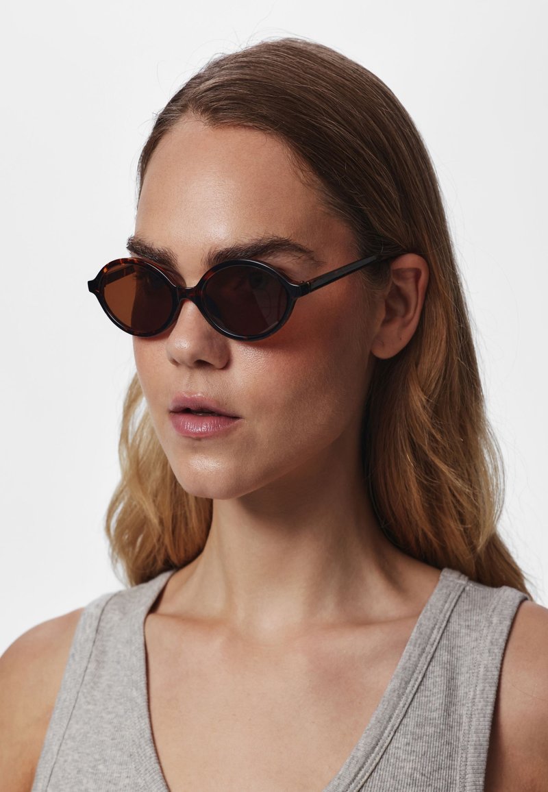 Femme aux longs cheveux châtain clair portant des lunettes de soleil rondes foncées et un haut gris sans manches sur un fond uni.