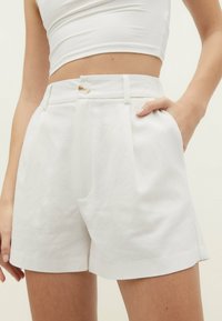 Pantaloni corti bianchi a vita alta con pieghe frontali, tasche laterali e chiusura con bottone. Tessuto testurizzato con un motivo sottile.