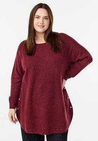 Sieviete, kura ģērbusies brīvā formas bordo džemperī ar sāniem uz pogām, stāv ar vienu roku uz gurniem pret baltu fona.