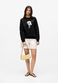 Bluză neagră cu imprimeu cu personaj de desene animate, pantaloni scurți crem, geantă de umăr galbenă și șlapi negri. Design simplu și culori contrastante.