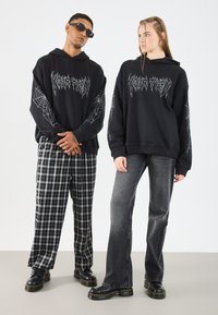 Svarte oversized hettegensere har edderkoppnett grafikk på ermene. Modell til venstre i svart rutete bukser, til høyre i grå jeans og svarte støvler.