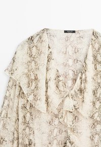 Genomskinlig blus med mönster av ormar i beige och brunt, med en volangkrage och mjuk tygtextur.