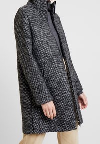 Cappotto in misto lana con colletto alto, motivo grigio-nero testurizzato, chiusura con zip e tasche laterali. Design minimale con taglio dritto.