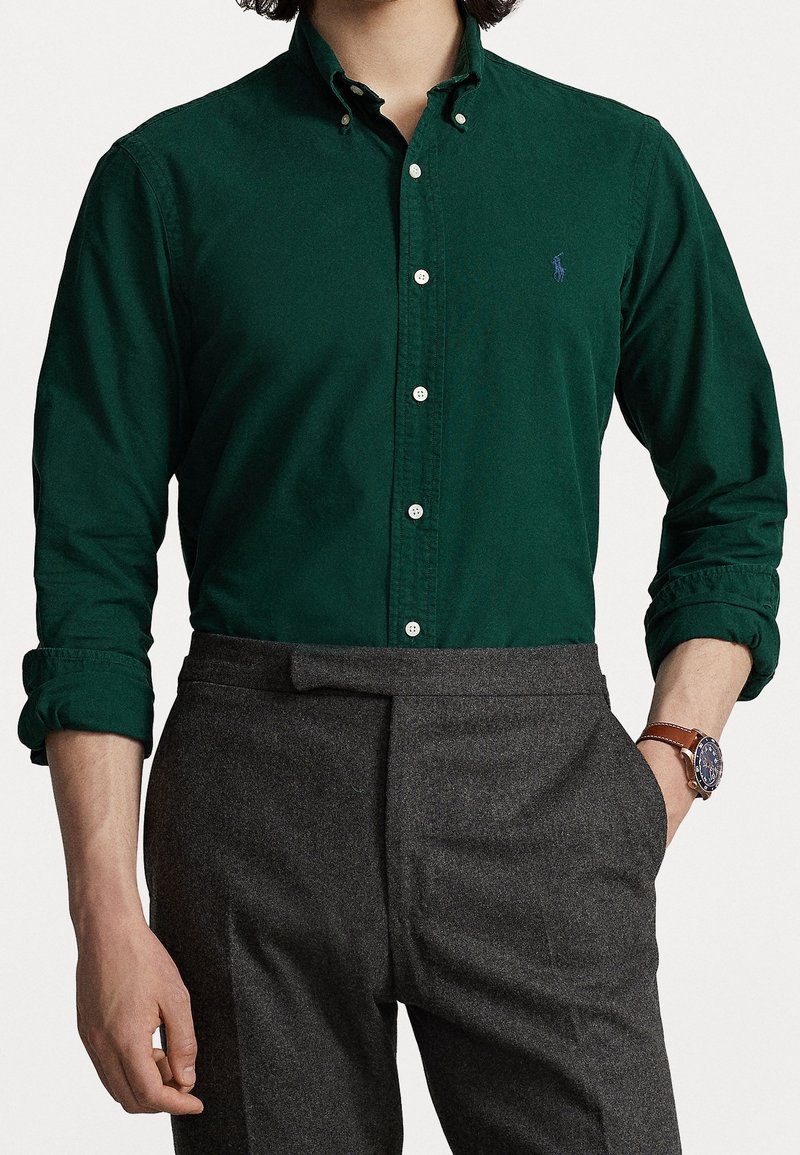 Ciemnozielona koszula zapinana na guziki z gładkiej tkaniny z klasycznym kołnierzykiem, zestawiona z szarymi, dopasowanymi spodniami i zegarkiem z brązowym paskiem ze skóry.