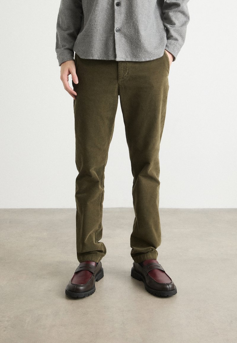 Pantalons en velours côtelé vert olive au design à jambes droites, associés à des mocassins bordeaux et noirs. Texture lisse avec des coutures subtiles.