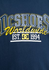 Námořnicky modré tričko s výrazným bílým a žlutým nápisem: "DCSHOES Worldwide EST. 1994" s texturovanou tkaninou a ležérním designem.