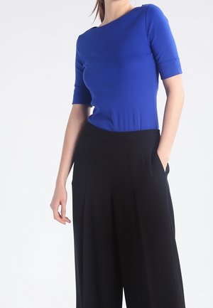 Femme portant un haut bleu ajusté à manches courtes, rentré dans un pantalon noir taille haute à jambes larges, la main dans la poche.