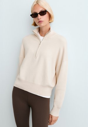 Vrouw met kort blond haar die zwarte rechthoekige zonnebrillen draagt, een crèmekleurige trui met halve rits, wit overhemd en donkerbruine broek, staand tegen een lichte muur.