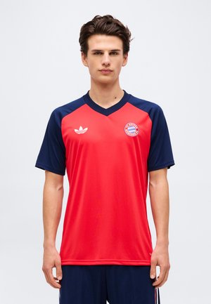 Giovane uomo che indossa la maglia da calcio rosso e blu navy del FC Bayern Monaco con i loghi Adidas, in piedi davanti a uno sfondo bianco neutro.