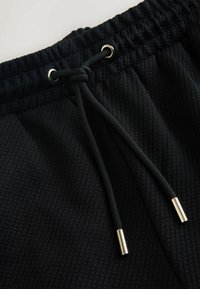 Shorts en tissu texturé noir avec taille élastique et cordons noirs terminés par des embouts métalliques argentés.