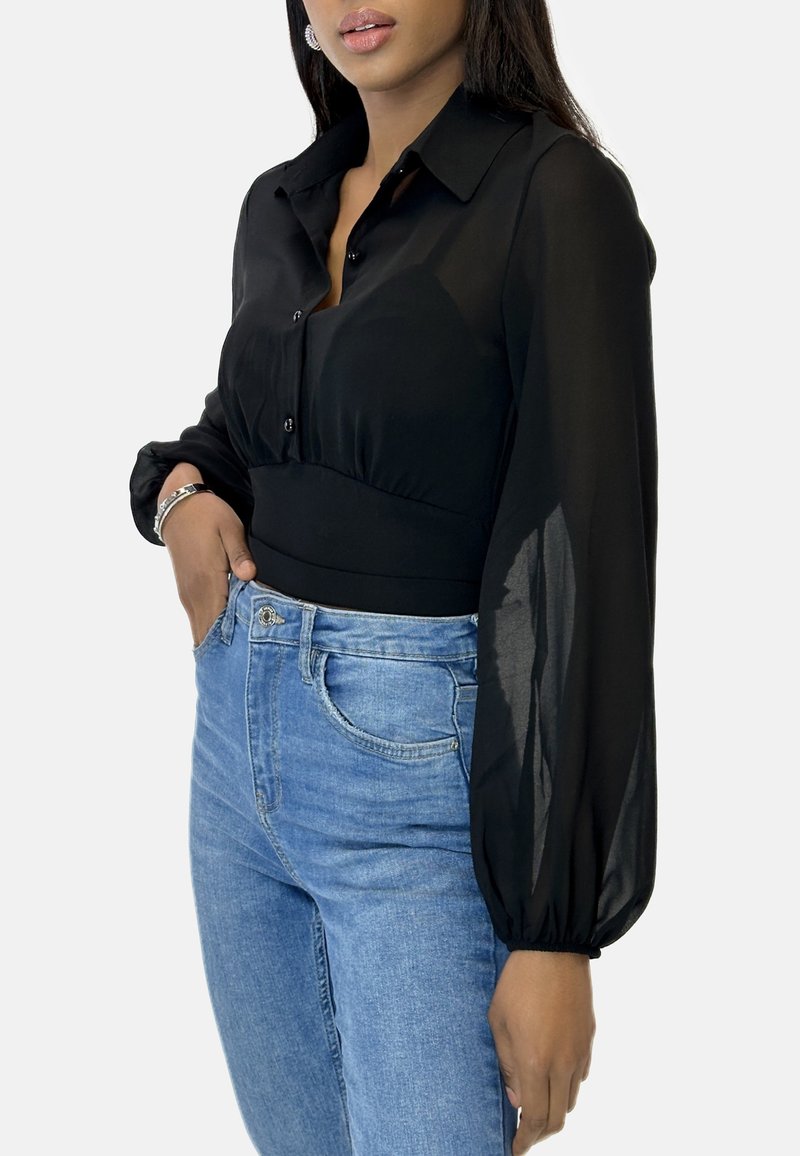 Blusa nera a maniche lunghe con corpetto aderente, chiusura frontale con bottoni e tessuto trasparente. Abbinata a jeans azzurri a vita alta.