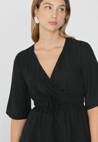 Schwarzes Wickelkleid mit kurzen Ärmeln, strukturiertem Stoff und tailliertem Bindegürtel. Hat einen tiefen V-Ausschnitt und dezente vertikale Rillen.
