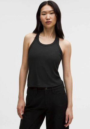 Frau mit langen dunklen Haaren, die ein ärmelloses schwarzes, geripptes Oberteil und schwarze Hosen trägt, steht vor einem schlichten hellen Hintergrund.