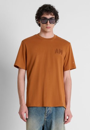 Joven con cabello rizado que lleva grandes gafas de sol negras, una camiseta marrón con "AM" en el pecho y vaqueros azules desteñidos frente a un fondo liso.