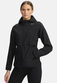 Giacca antivento nera con zip frontale, cappuccio, polsini elastici e due tasche laterali. Presenta un logo visibile sulla parte superiore sinistra del petto.