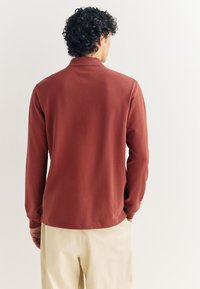 Polo de manga larga de color óxido, hecho de tela suave, con cuello plano y aberturas laterales, combinado con pantalones de color claro.