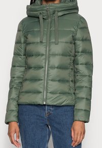 Giacca verde imbottita con zip frontale, cappuccio regolabile con cordino e dettagli a costine, caratterizzata da una texture trapuntata e un design aderente.