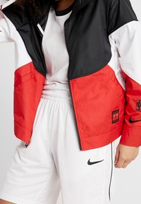 Färgsplittad jacka med svarta, röda och vita paneler. Tillverkad av slät tyg, har dragkedjestängning och NBA-logga på ärmen. Vita shorts nedanför.