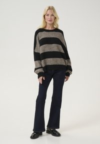 Maglione a righe in nero e beige, design oversize con spalle scese, abbinato a jeans scuri a zampa d'elefante e scarpe nere.