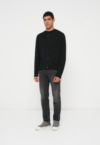 Must kardigan nuppude kinnitusega, kantud musta särgi peal. Sobitatud hallide teksade ja roheliste tossudega, seismas valge tausta ees.