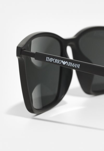Ochelari de soare negri cu un design îndrăzneț și unghiular. Lentilele sunt întunecate și reflectorizante. Brațele cu temple au logo-ul "Emporio Armani" în argintiu.