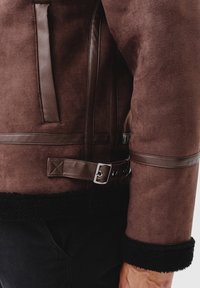 Giacca in finta pelle scamosciata marrone con dettagli in pelle marrone, tasca laterale, dettagli con fibbia regolabile e rivestimento in shearling nero ai polsini.
