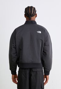 Veste bomber noire avec un col et des poignets côtelés, affichant un logo blanc dans le dos. Le tissu paraît lisse et isolant.