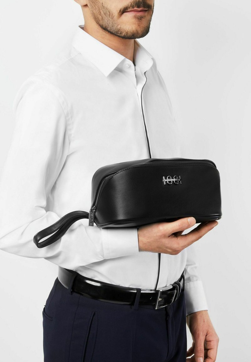 Homme en chemise blanche tenant une trousse de toilette en cuir noir avec fermeture éclair et bracelet contre un fond uni.