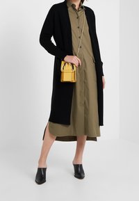 Svart stickad kofta över en olivgrön skjortklänning med sidospalter. Liten gul handväska med guld detaljer. Svarta mular.