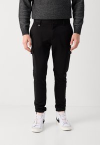 Černé cargo kalhoty s úzkým střihem, vybavené elegantními bočními kapsami, hladkou texturou látky a knoflíkovým zapínáním v pase. Kombinovány s bílými teniskami.