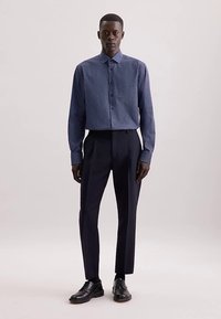 Donkerblauw overhemd met knopen en witte polka dots, lange mouwen en een kraag, gecombineerd met op maat gemaakte zwarte broek en zwarte nette schoenen.