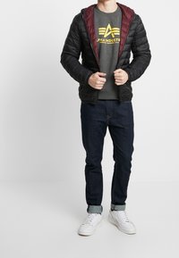 Giacca a piumino nera con fodera color amaranto, felpa grigia con logo giallo, jeans blu scuro e scarpe da ginnastica bianche. La modella è in piedi con le mani sulla giacca.